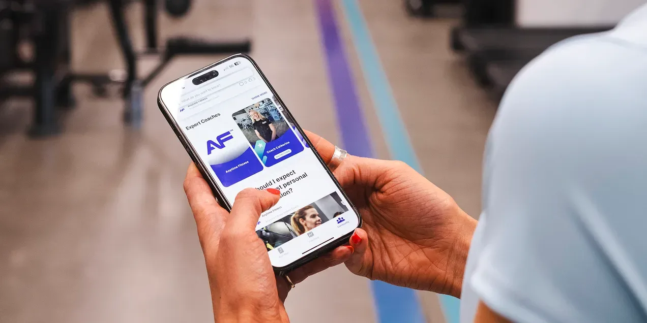 A person holds a smartphone displaying the Anytime Fitness website, featuring options for expert coaching and fitness information.